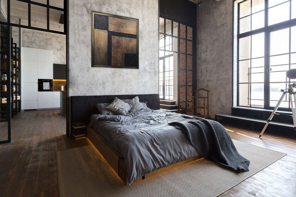 Blue Copper Wash by Alli Cullimore displayed above a bed in a moody industrial loft bedroom — a square abstract painting with dark blue and copper tones in a dark bronze frame, Gallery 68